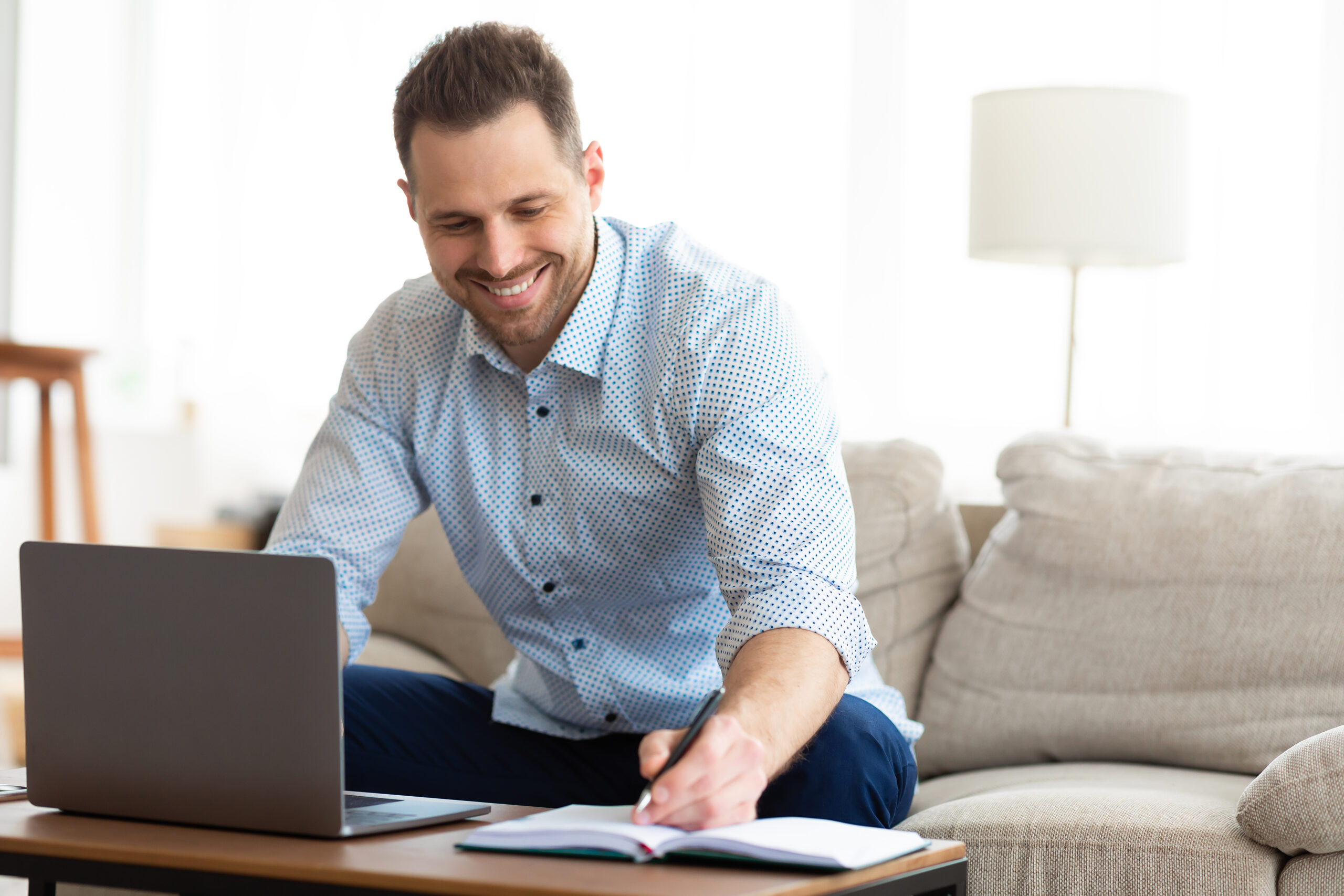 Note Taking Concept. Smiling handsome man using laptop, writing in notebook, sitting on couch, copy space